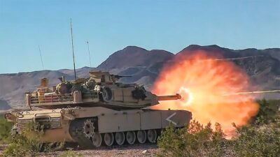 A tank fires a round in a desert landscape with mountains in the background, emitting a burst of flame from its cannon.