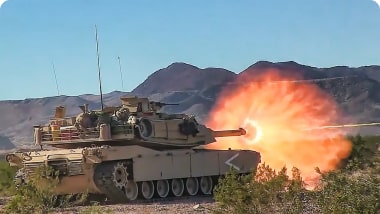 A tank firing its main cannon in a desert landscape, with mountains in the background and flames visible from the barrel.